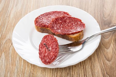 Sandwich with smoked sausage and piece of sausage strung on fork in plate on wooden tableの写真素材