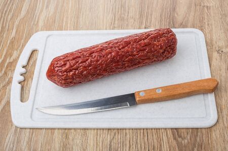 Smoked sausage and kitchen knife on plastic cutting board on on wooden tableの写真素材