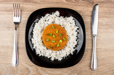 Black plate with cutlet and boiled white rice, fork and knife on wooden table. Top viewの写真素材