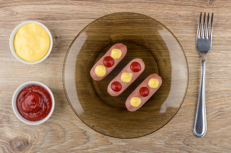 Small boiled sausages with sauces in brown plate, bowls with ketchup and mayonnaise on wooden table. Top viewの写真素材