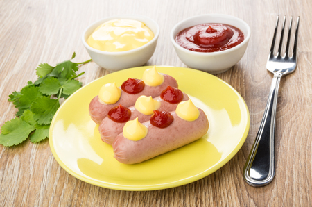 Three small boiled sausages with ketchup and mayonnaise in yellow plate, cilantro and fork on wooden tableの写真素材