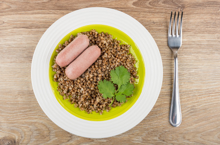 Boiled buckwheat with sausages in plate and fork on wooden table. Top viewの写真素材