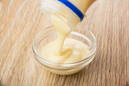 Pouring sweet condensed milk from bottle in transparent bowl on wooden tableの写真素材