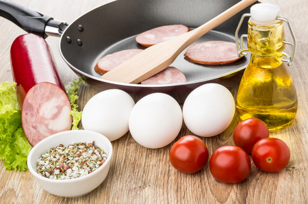 Slices of smoked sausage in frying pan, eggs, vegetable oil, spices and tomatoes on wooden tableの写真素材