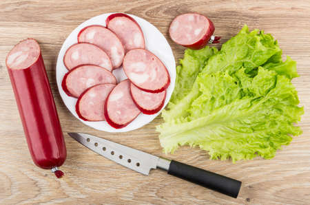 Slices of smoked sausage in white plate, leaves of lettuce and knife on wooden table. Top viewの写真素材