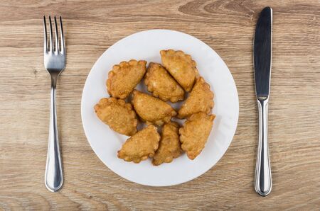 Small fried chebureks in white glass plate, knife and fork on wooden table. Top view