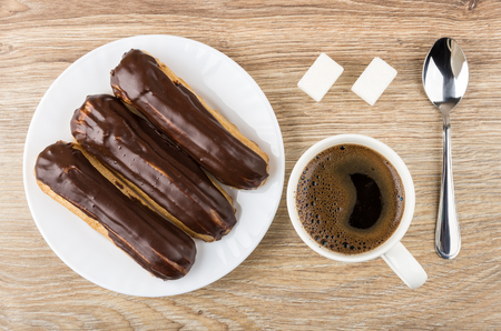 Eclairs with chocolate in plate, coffee in cup, lumpy sugar and spoon on wooden table. Top view