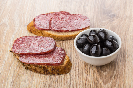 Sandwiches with smoked sausage and bowl with black olives on wooden tableの写真素材