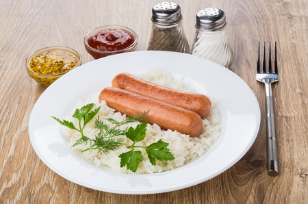 Fried sausages with boiled rice, bowls with mustard and ketchup, salt, pepper and fork on wooden tableの写真素材