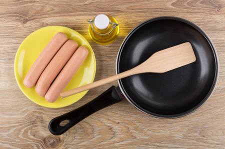 Raw peeled sausages in saucer, bottle of vegetable oil, frying pan and spatula on wooden table. Top view on wooden tableの写真素材