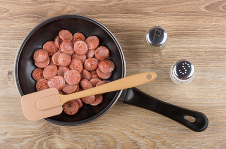 Pieces of fried sausage in frying pan, spatula, salt and pepper on wooden table. Top viewの写真素材