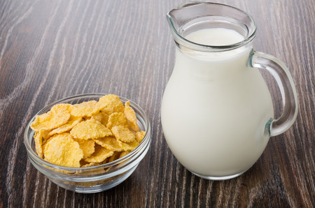 Transparent bowl with corn flakes and jug of milk on dark wooden table