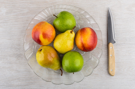 Nectarines, pears, apples in transparent dish and kitchen knife on wooden table. Top viewの写真素材
