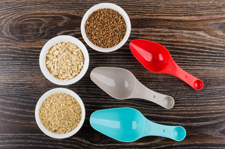 Bowls with oat flakes, brown rice and buckwheat and plastic spoons on wooden table. Top viewの写真素材
