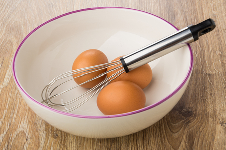 Whisk and chicken eggs in ceramic bowl on wooden tableの写真素材