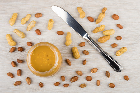 Opened jar with peanut butter, knife and scattered peanuts on wooden table. Top view