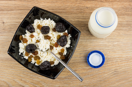 Cottage cheese with raisins and prunes and teaspoon in black bowl, open bottle of yogurt on wooden table. Top viewの写真素材