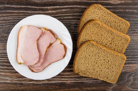 Slices of smoked gammon in plate, rye bread on wooden table. Top viewの写真素材