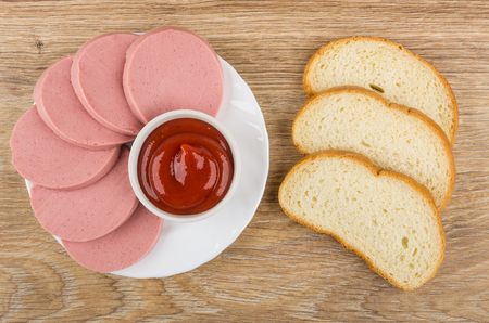 Slices of sausage and ketchup in plate, pieces of bread on wooden table. Top viewの写真素材
