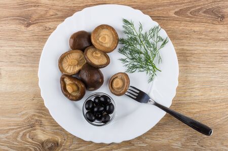 Marinated mushrooms, black olives and dill, fork in dish on wooden table. Top view