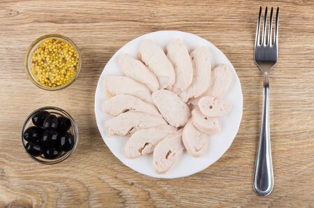 Slices chicken breast in plate, fork, mustard and black olives on wooden table. Top viewの写真素材