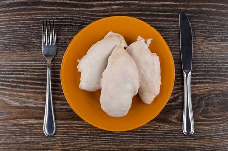 Pieces of boiled chicken breast in orange plate, fork and knife on wooden table. Top viewの写真素材