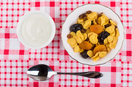Corn flakes with dried fruits in bowl, sour cream and spoon on table. Top view