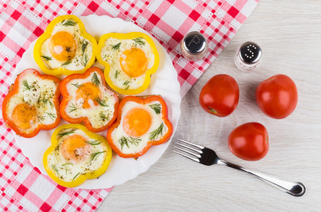 Fried eggs with red and yellow sweet peppers in dish, condiments, tomatoes and fork on wooden table....