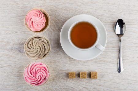 Three cupcakes with cream, teaspoon, cup of tea, lumpy sugar on wooden table. Top viewの写真素材