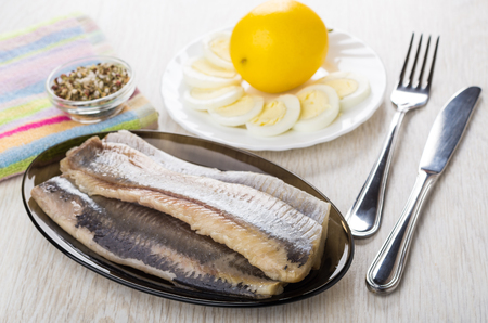 Fillet of herring in dish, lemon, boiled eggs in plate, condiment, knife and fork on wooden tableの写真素材