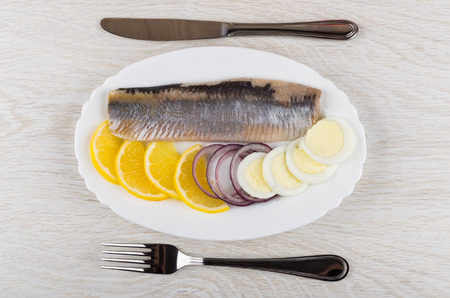 Fillet of herring with lemon, onion, egg in dish, knife and fork on wooden table. Top viewの写真素材