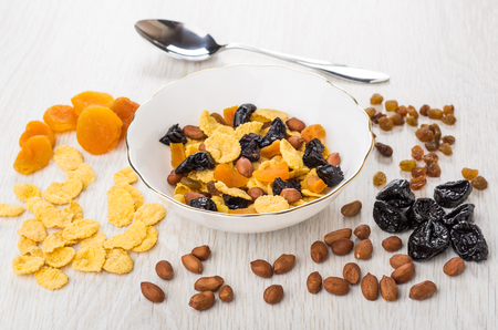 Muesli in bowl, scattered dried fruits, corn flakes and nuts, spoon on wooden tableの写真素材