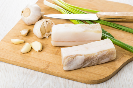 Pieces of salted lard, garlic and green onion on cutting board on wooden tableの写真素材