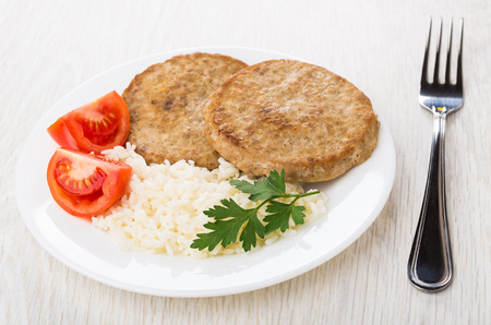 Fried cutlets in plate with rice, pieces of tomato, parsley and fork on wooden tableの写真素材