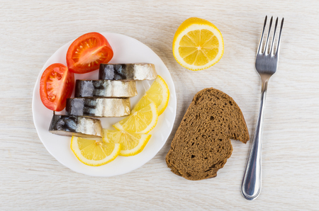 Plate with slices of smoked mackerel, lemon, tomato, piece of bread and fork on wooden table. Top viewの写真素材