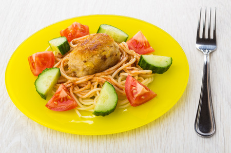 Spaghetti with fried cutlet, cucumbers, tomato in yellow plate and fork on wooden tableの写真素材