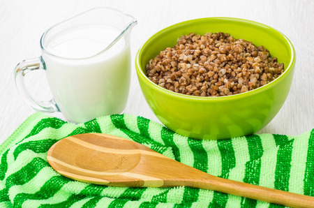 Boiled buckwheat in green bowl, jug of milk, striped napkin and bamboo spoon on wooden tableの写真素材