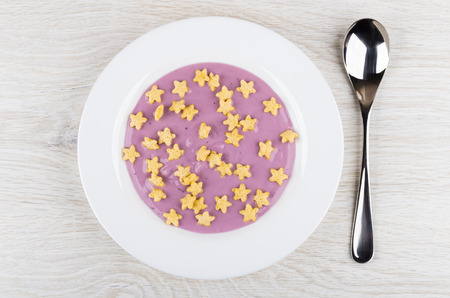 Blueberry yogurt with cornflakes in form star in white plate, spoon on wooden table. Top view