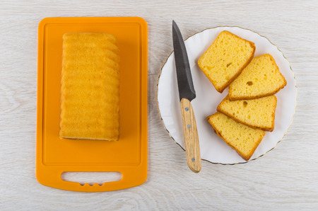 Piece of biscuit  on cutting board and slices of biscuit, knife in plate on wooden table. Top view