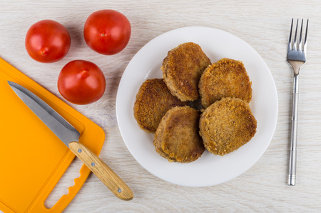 Fried meat cutlets in plate, tomatoes, cutting board, knife and fork on wooden table. Top view