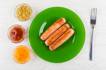 Fried sausages in green plate, bowls with ketchup, mustard, fork on wooden table. Top viewの写真素材