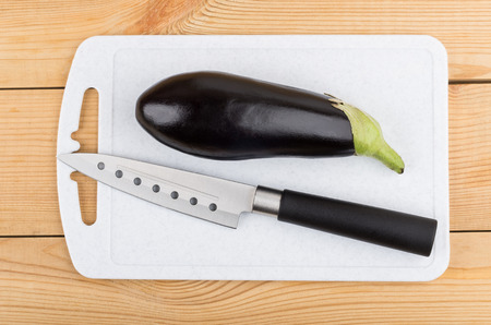 Whole eggplant and kitchen knife on plastic cutting board on wooden table. Top viewの写真素材