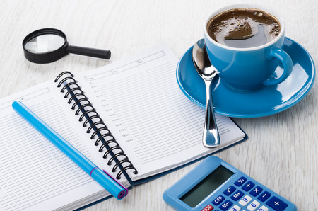 Calculator, notepad, pen, magnifying glass, coffee in blue cup on saucer on wooden tableの写真素材
