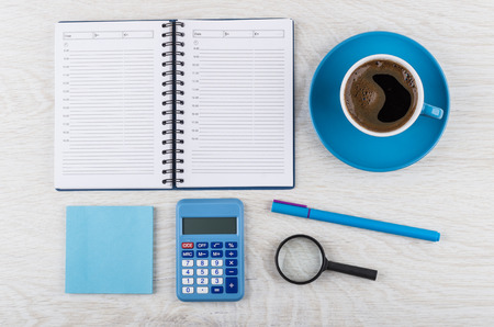 Opened notepad, coffee in blue cup on saucer, block of paper, calculator, pen and magnifying glass on wooden table. Top viewの写真素材