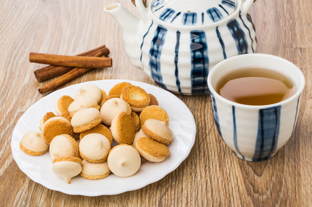Small cookies with egg glaze on plate, teapot, cinnamon sticks, cup of tea on wooden tableの写真素材
