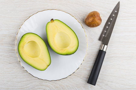 Two halves of avocado in white plate, nucleus, kitchen knife on wooden table. Top viewの写真素材