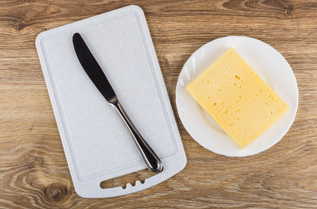 Piece of cheese in plate, table knife on plastic cutting board on wooden table. Top viewの写真素材