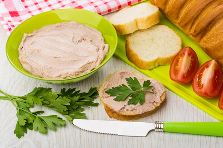 Pate in green bowl, parsley, sandwich with liver pate, pieces of tomato, loaf of bread on cutting board, knife on wooden tableの写真素材
