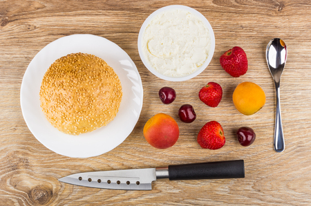 Bun with sesame, cream cheese in jar, kitchen knife, spoon, apricots, cherries and strawberries on wooden table. Top viewの写真素材