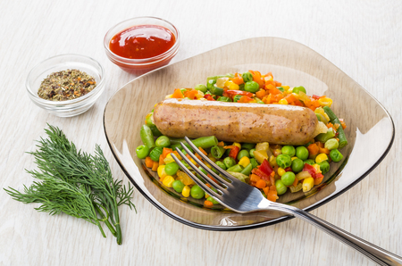 Fried sausage with vegetable mix in plate, bowls with condiment and ketchup, dill, fork on wooden tableの写真素材